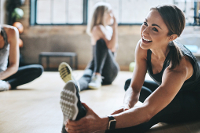 You can become a trainer (or hire a trainer) at Dash Class. Three women stretching in a gym. The most prominent lady is smiling while doing a sitting single leg hamstring stretch.